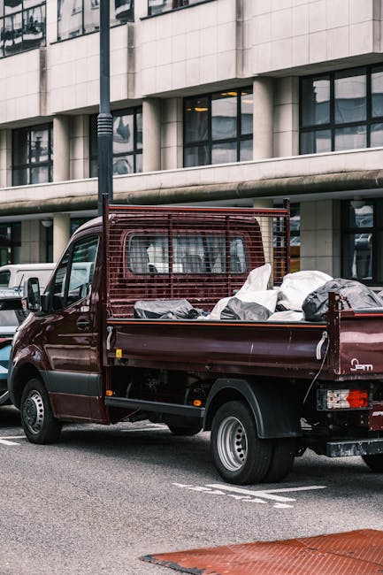 A small, dark red flatbed pickup truck parked on a city street in front of a modern office building with large glass windows and a white stone facade. The truck's cargo bed is filled with various type
