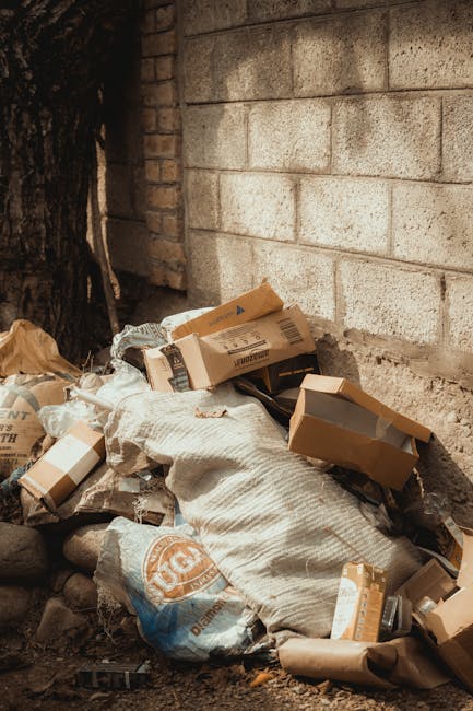 Two large black garbage bags and a cardboard box, all filled with waste, are positioned on the pavement near a black metal fence and a leafy shrub in an outdoor urban setting. The bags appear to be ti